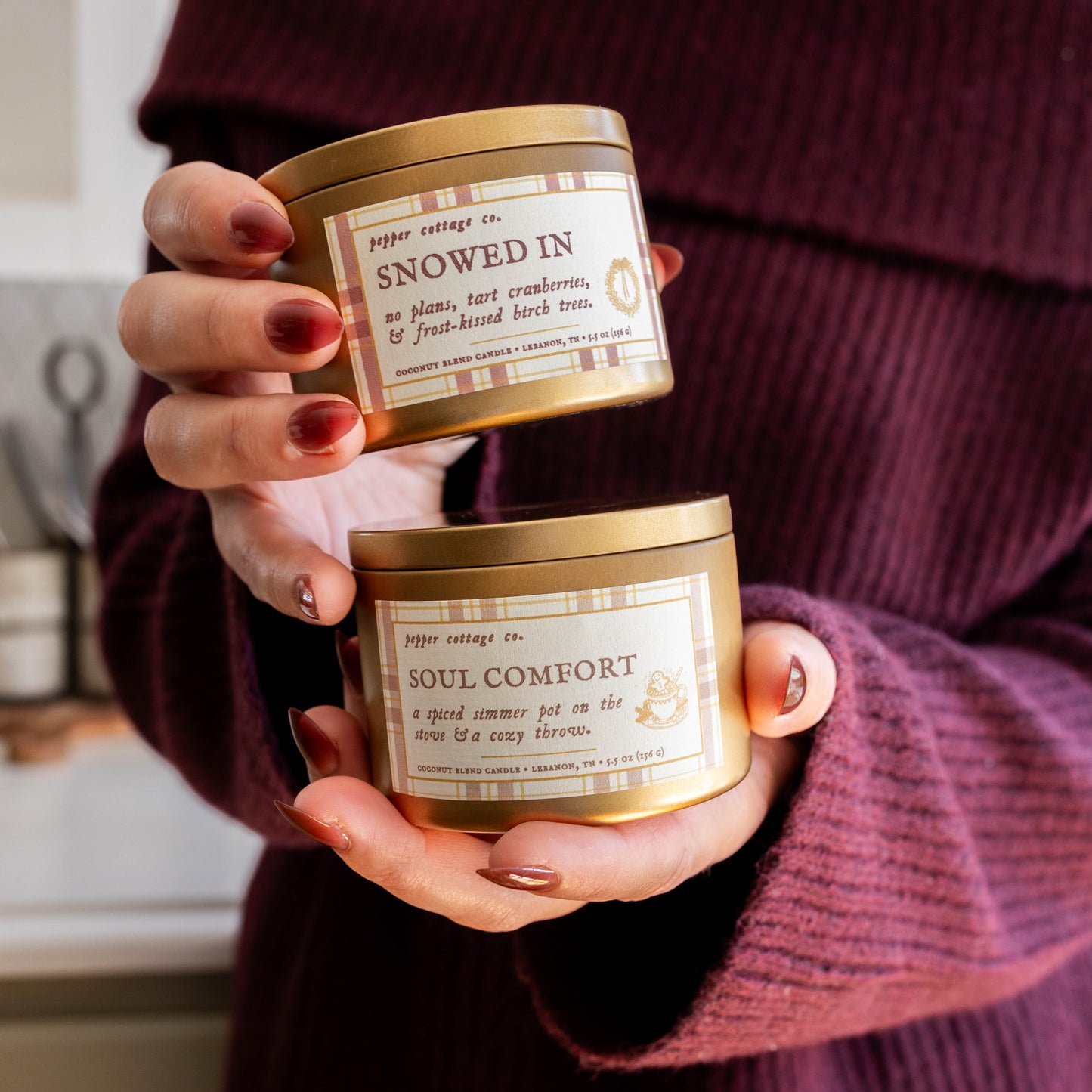 Person holding two candles labeled 'Snowed In' and 'Soul Comfort' in a home setting.